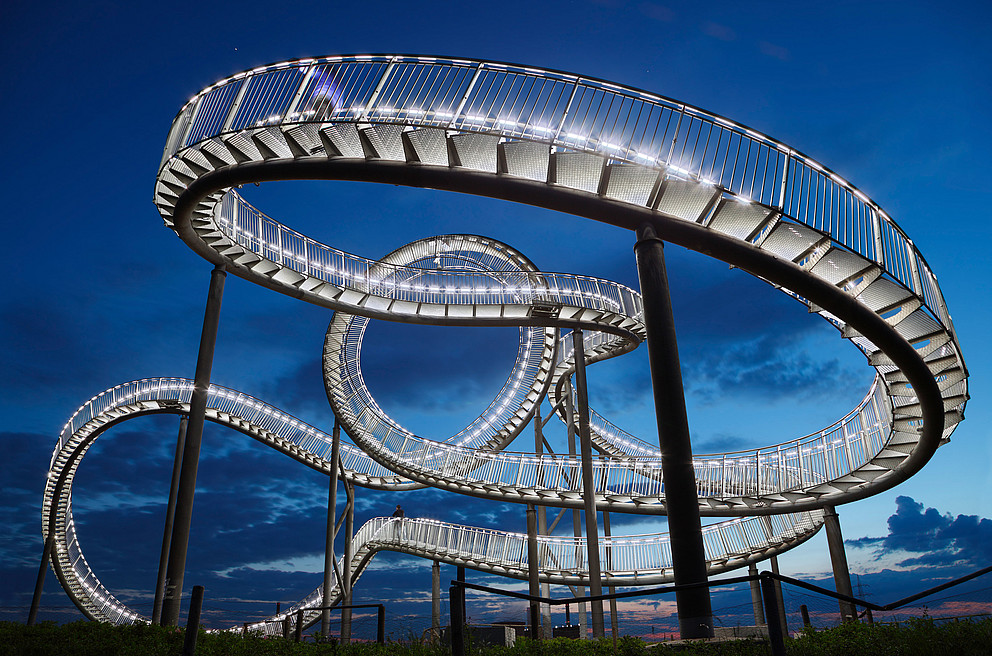 Illuminated staircase sculpture “Tiger and Turtle,” designed to resemble a roller coaster, against the evening sky